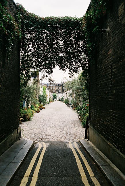 A narrow cobblestone street viewed through an archway formed by dense, green foliage and climbing plants, creating a natural canopy overhead. The street is lined with potted plants and small trees alo