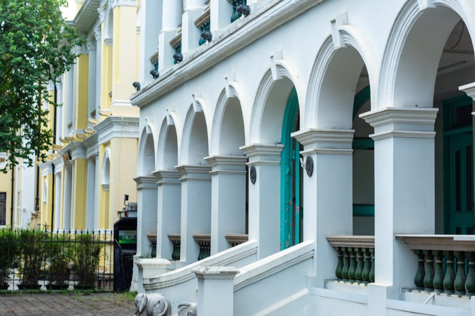 A row of elegant terraced houses with arched doorways supported by white columns, featuring vibrant blue and green doors. The facades of the buildings are painted in pastel shades of yellow and white,